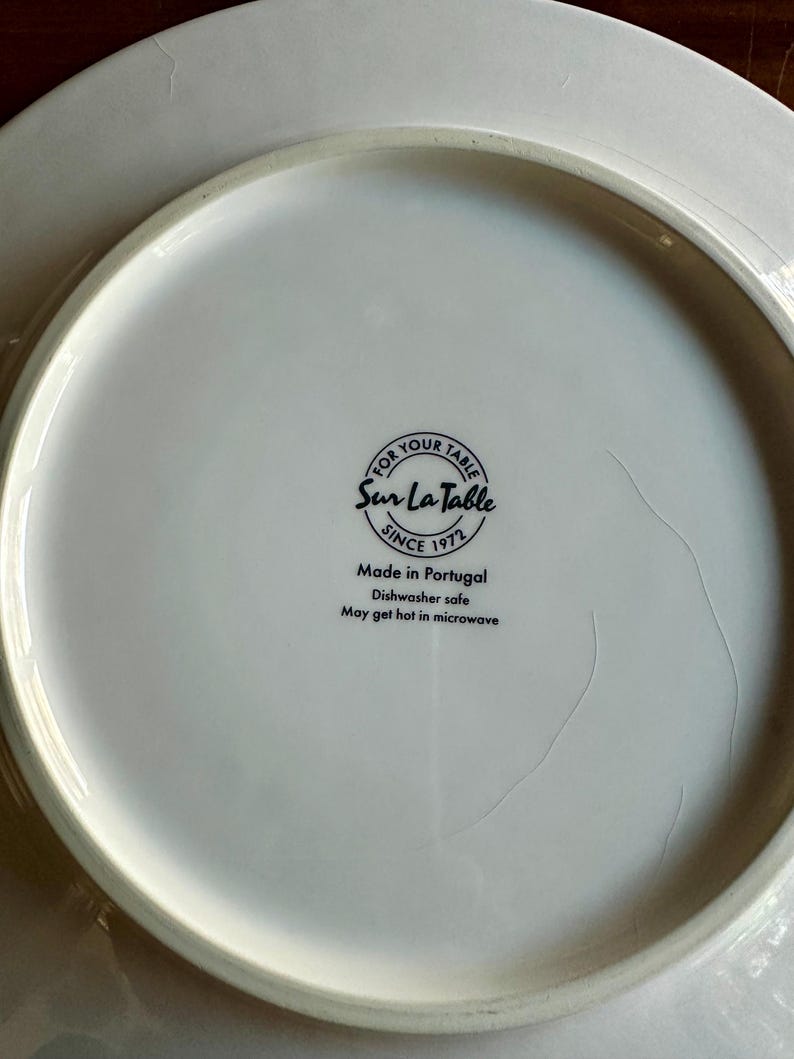 Peut inclure: Assiette en c&eacute;ramique blanche avec le texte "FOR YOUR TABLE Sur La Table SINCE 1972 Made in Portugal Dishwasher safe May get hot in microwave". L'assiette est de forme circulaire.