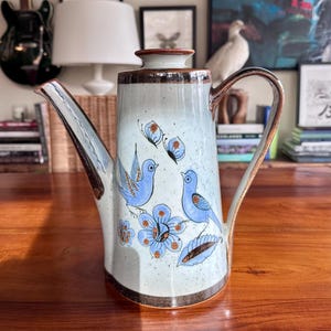 May include: A ceramic teapot with a light blue background, featuring blue birds, butterflies, and flowers. The teapot has a brown rim, handle, and spout. The teapot is sitting on a wooden table.