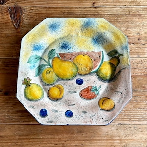 May include: A ceramic plate with a yellow, blue, and white background featuring a painted design of fruit, including a watermelon, pears, lemons, and blueberries.
