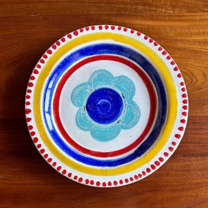 May include: A round ceramic plate featuring a white center with a central dark blue circle surrounded by a light blue flower petal design. The plate is decorated with concentric rings in red, dark blue, and yellow, finished with a red dotted rim.