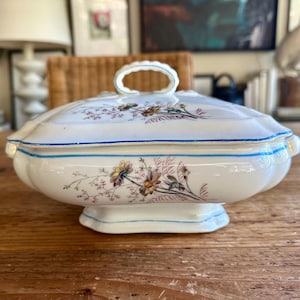 May include: A white porcelain serving dish with a lid, featuring a floral design in yellow, brown, and purple. The dish has a blue trim and a handle on the lid. It sits on a wooden surface.