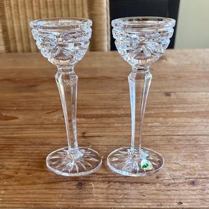 May include: A pair of clear glass candle holders with a cut glass design. The candle holders have a tall, slender stem and a round base.