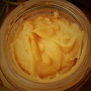 May include: A close-up of a glass jar filled with a thick, golden yellow substance. The substance has a smooth, creamy texture and appears to be a type of honey or syrup.