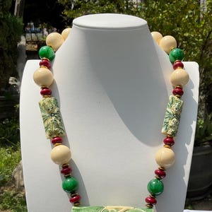 May include: A white and green carved bone necklace with red beads and a large pendant featuring three frogs. The pendant is carved with intricate details and shows the frogs sitting on a leaf.