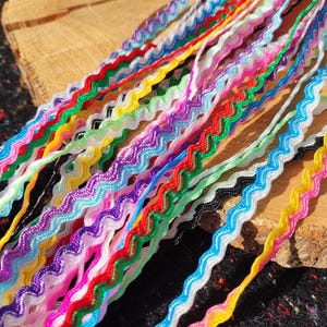 Op de afbeelding: Een verzameling van meerdere strengen zigzagband in een verscheidenheid aan levendige kleuren, waaronder rood, geel, groen, blauw, paars, roze en wit. Elk afzonderlijk lint heeft een consistent golvend zigzagpatroon over de gehele lengte.