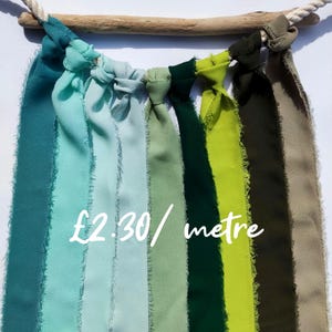 May include: A decorative wall hanging featuring strips of fabric in shades of teal, green, and olive green, knotted and suspended from a wooden dowel. The fabric strips have a frayed edge. A rope is attached to the wooden dowel.