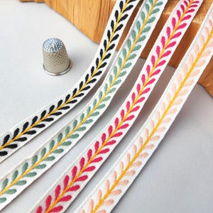 Könnte beinhalten: Vier Streifen aus weißem Webband mit einem gelben Mittelsteg und gestickten Blättern in Schwarz, Salbeigrün, leuchtendem Pink und hellem Pfirsich. Ein kleiner, metallischer Fingerhut mit Noppen liegt links neben den Bändern auf einer Fläche.