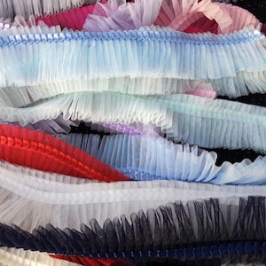 May include: A close-up of layered, ruffled trim in various colors. The trim includes shades of blue, white, red, pink, and black. The fabric appears to be a sheer, lightweight material, possibly tulle or netting, with a pleated or gathered design.