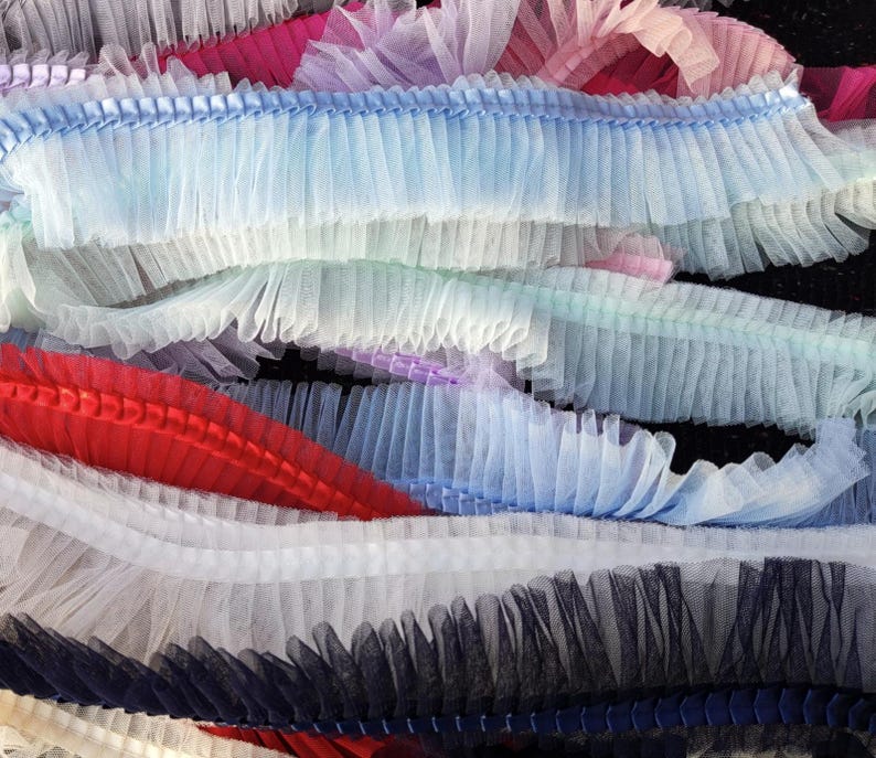 May include: A close-up of layered, ruffled trim in various colors. The trim includes shades of blue, white, red, pink, and black. The fabric appears to be a sheer, lightweight material, possibly tulle or netting, with a pleated or gathered design.
