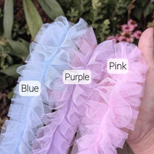 Pu&ograve; includere: Tre strisce di tessuto in tulle arricciato nei colori azzurro, viola chiaro e rosa chiaro. Ogni striscia presenta una trama a rete stratificata e arricciata. Etichette bianche con testo nero identificano le strisce come Blue, Purple e Pink.