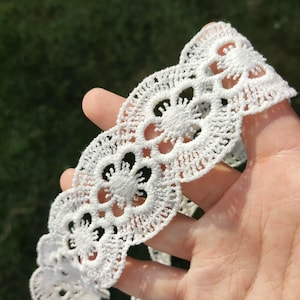 May include: Close-up of white lace trim featuring a repeating floral design. The lace is held in a hand, highlighting its delicate openwork pattern. The trim is made of a soft material, possibly cotton.