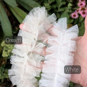 Pu&ograve; includere: Due strisce di nastro in tulle arricciato, una color crema tenue e una color bianco brillante. Ogni striscia presenta una trama a rete increspata e stratificata con un motivo a griglia fine e semitrasparente. Testo Cream e White visibile.