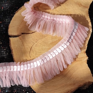 May include: Pink ruffled trim with a sheer, layered design. The trim features a satin ribbon down the center and is displayed on a wooden surface. The word "Pink" is printed in a bold, white font at the bottom.