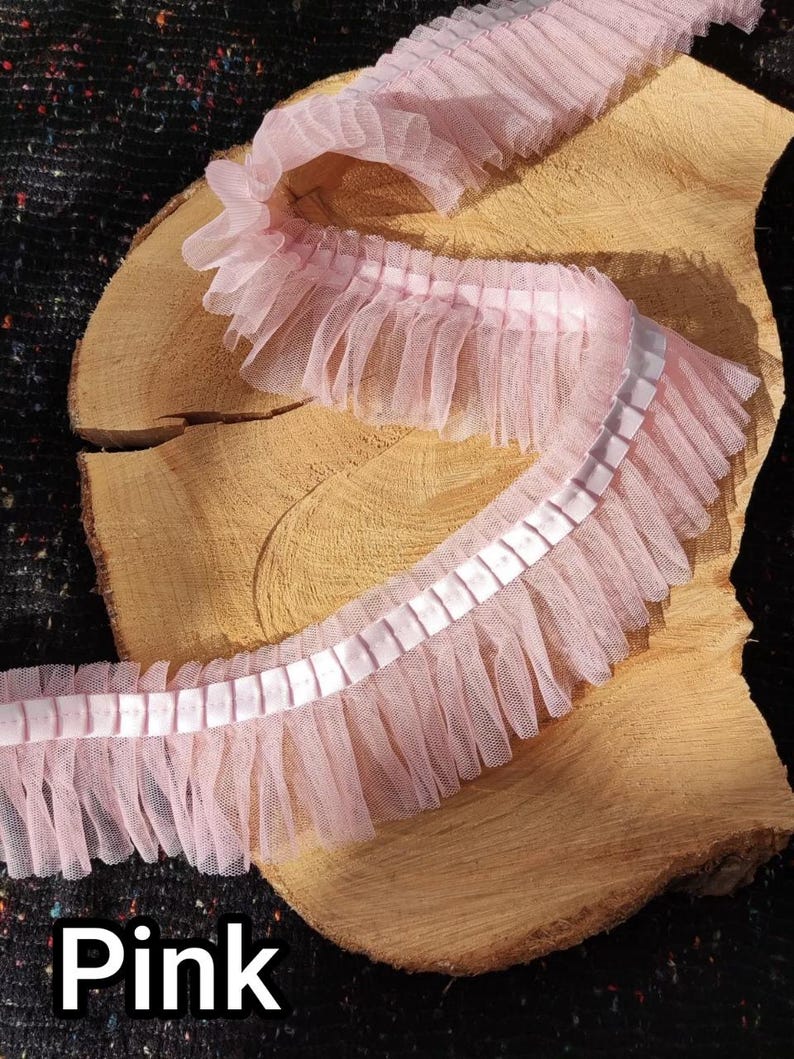 May include: Pink ruffled trim with a sheer, layered design. The trim features a satin ribbon down the center and is displayed on a wooden surface. The word "Pink" is printed in a bold, white font at the bottom.
