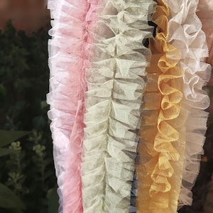 Pu&ograve; includere: Una collezione di quattro strisce di tessuto in tulle arricciato disposte verticalmente. I colori includono un bianco tenue, un rosa pallido, un verde salvia tenue e un giallo oro caldo. Ogni striscia presenta una trama a rete fine.
