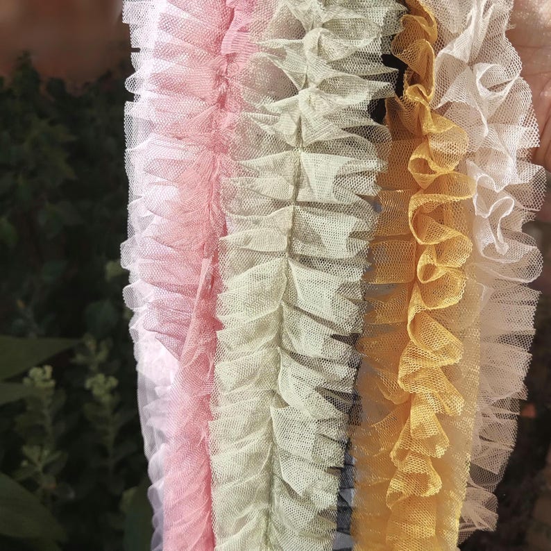 Pu&ograve; includere: Una collezione di quattro strisce di tessuto in tulle arricciato disposte verticalmente. I colori includono un bianco tenue, un rosa pallido, un verde salvia tenue e un giallo oro caldo. Ogni striscia presenta una trama a rete fine.