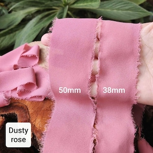 Puede incluir: Dos tiras de cinta de tela color rosa polvoriento con bordes deshilachados y sin rematar. Las cintas tienen texto que indica anchos de 50mm y 38mm. El material tiene una apariencia mate y texturizada, mostrada en una longitud continua.