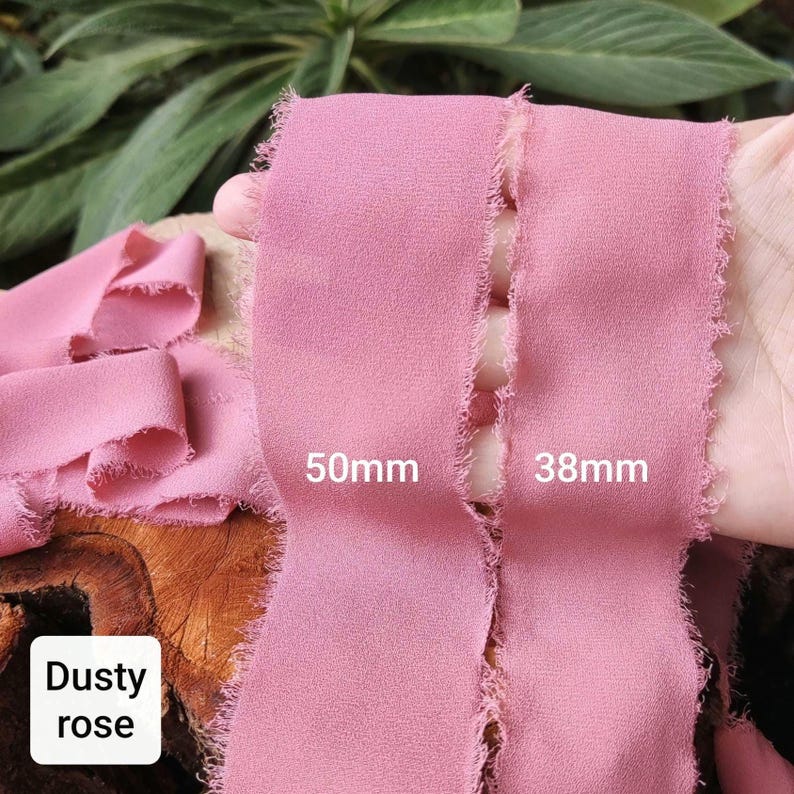 Puede incluir: Dos tiras de cinta de tela color rosa polvoriento con bordes deshilachados y sin rematar. Las cintas tienen texto que indica anchos de 50mm y 38mm. El material tiene una apariencia mate y texturizada, mostrada en una longitud continua.