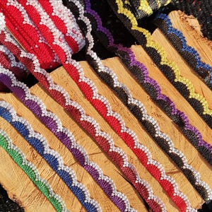 Può includere: Una collezione di nastri decorativi colorati. I nastri sono in vari colori, tra cui rosso, viola, blu, verde e nero. Ogni nastro ha un bordo smerlato con un bordo bianco a contrasto. I nastri sono disposti su una superficie di legno.