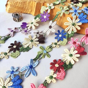 Può includere: Un bordo decorativo con un motivo continuo di fiori ricamati in vari colori, tra cui rosso, blu, viola, rosa, bianco e verde. Il nastro di pizzo floreale è disposto in più fili, mostrando cuciture intricate e dettagli a forma di foglia.