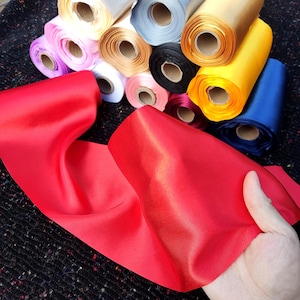 May include: A collection of satin ribbon rolls in various colours including red, yellow, blue, black, white, pink, and gold. The rolls are arranged in a cluster, with a long, smooth, vibrant red satin ribbon draped in the foreground.
