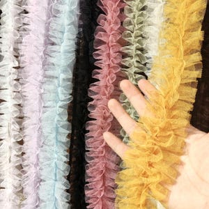 Pu&ograve; includere: Una collezione di strisce di passamaneria in tulle arricciato disposte verticalmente in vari colori, tra cui bianco, viola chiaro, azzurro, nero, rosa antico, verde salvia e giallo senape, con una trama a rete fine e bordi arricciati.