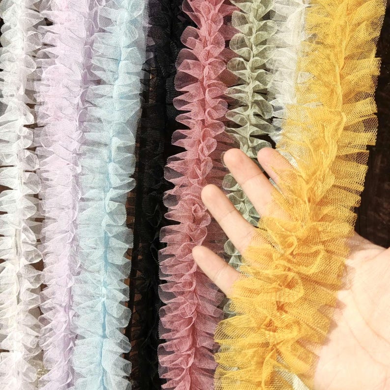 Pu&ograve; includere: Una collezione di strisce di passamaneria in tulle arricciato disposte verticalmente in vari colori, tra cui bianco, viola chiaro, azzurro, nero, rosa antico, verde salvia e giallo senape, con una trama a rete fine e bordi arricciati.