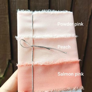 Puede incluir: Tres cintas de tela apiladas con bordes deshilachados, atadas con un fino cord&oacute;n met&aacute;lico plateado. Las cintas est&aacute;n etiquetadas como Powder pink, Peach y Salmon pink, mostrando un degradado de texturas de tela en tonos rosa y naranja.