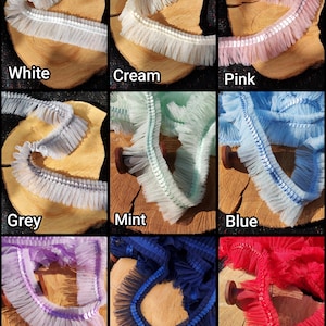 May include: A collage of colorful ruffled trim samples. Colors include white, cream, pink, grey, mint, blue, purple, navy, and red. Each trim is displayed on a wooden slice, showcasing the texture and variety of the fabric.