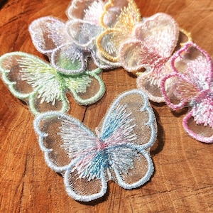 May include: A collection of embroidered butterfly patches featuring sheer mesh wings with iridescent thread detailing in pastel shades of pink, blue, yellow, and green. Each butterfly has a distinct colour outline and intricate stitching on the wings.