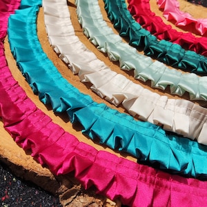 Könnte beinhalten: Eine Sammlung farbenfroher Satinbänder mit Rüschen in den Farbtönen Fuchsia, Türkis, Weiß, Mintgrün, Smaragdgrün und Rot. Die Bänder sind kreisförmig auf einer Holzoberfläche angeordnet und zeigen ihre Textur und lebendigen Farben.