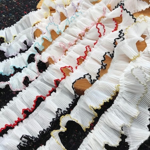 Könnte beinhalten: Nahaufnahme von mehreren Reihen weißer, plissierter Stoffborte mit farbigen Kanten. Die Borte hat Kanten in verschiedenen Farben, darunter Rot, Schwarz, Gold, Rosa, Blau und Gelb. Der Stoff hat ein strukturiertes, gerüschtes Aussehen.