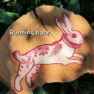 Könnte beinhalten: Ein gestickter Aufnäher eines laufenden Hasen mit roten Umrissen auf hellem Stoff. Der Hase zeigt einen Halskrausenkragen, florale Muster, kleine Sterne und das in Rot gestickte Wort "lived". Der Text "Running hare" ist oben zu sehen.