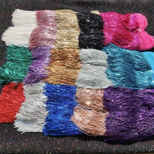 May include: A collection of fifteen rectangular fabric swatches featuring long, shimmering fringe trim. Each swatch displays a distinct solid colour, including white, pink, gold, black, magenta, green, purple, silver, teal, red, blue, and copper tones.