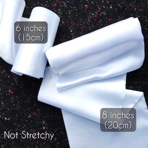 Puede incluir: Cinta de raso blanca, de 15 cm y 20 cm de ancho, se muestra sobre un fondo oscuro y moteado. También es visible el texto "Not Stretchy". La cinta está doblada y enrollada, mostrando su textura y brillo.