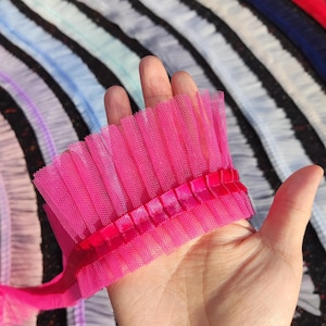 May include: A vibrant pink tulle trim with a pleated design, held in a hand. The trim features a satin ribbon detail. In the background, other colors of pleated tulle are visible, creating a colorful display of fabric.