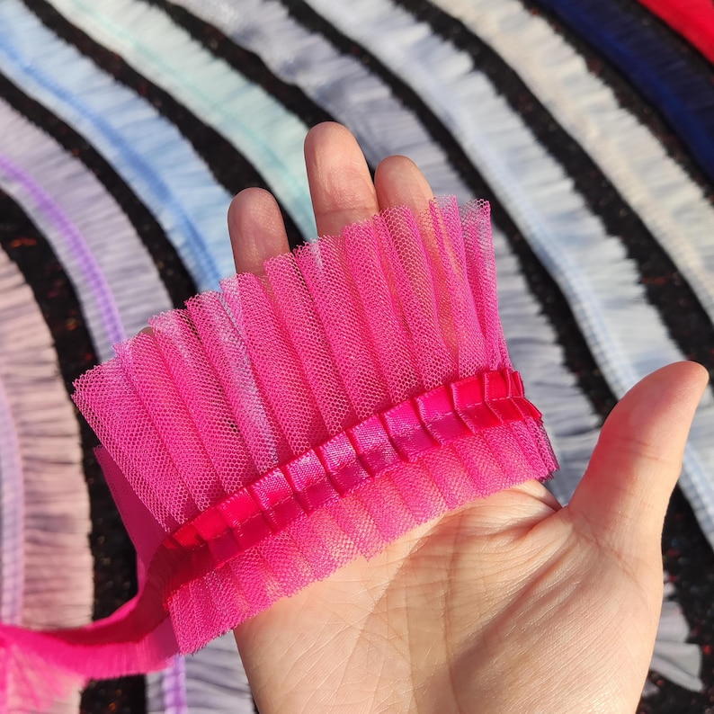 May include: A vibrant pink tulle trim with a pleated design, held in a hand. The trim features a satin ribbon detail. In the background, other colors of pleated tulle are visible, creating a colorful display of fabric.