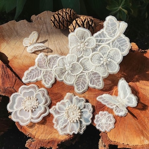 Könnte beinhalten: Eine Sammlung von cremeweißen bestickten Spitzenapplikationen mit Blumenmotiven und zwei Schmetterlingsformen. Das Set enthält ein großes Blumenarrangement, zwei einzelne Blüten, eine kleine Blume und zwei schmetterlingsförmige Spitzenteile.