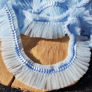 May include: Light blue tulle trim with a ruffled edge and a satin ribbon detail. The trim is arranged on a wooden surface, showcasing its delicate texture and color. This decorative trim is suitable for various craft projects.