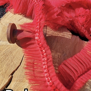 May include: Close-up of red tulle trim with a ruffled edge. The trim is layered and has a delicate, airy appearance. The word "Red" is displayed in a white font at the bottom left corner. The trim is likely used for crafting or sewing projects.