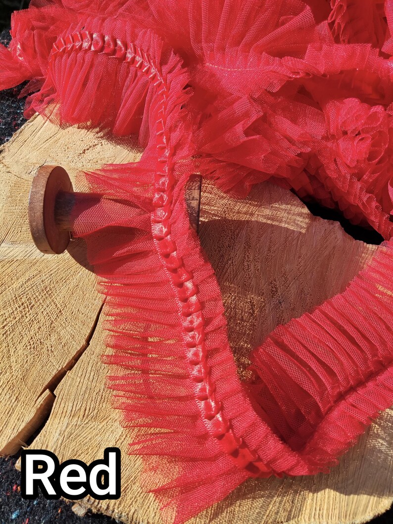 May include: Close-up of red tulle trim with a ruffled edge. The trim is layered and has a delicate, airy appearance. The word "Red" is displayed in a white font at the bottom left corner. The trim is likely used for crafting or sewing projects.