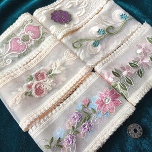 May include: A collection of five strips of sheer cream-coloured lace trim, each featuring distinct floral embroidery in shades of pink, purple, green, and blue. The edges are finished with scalloped detailing, and a small metal bobbin sits nearby.