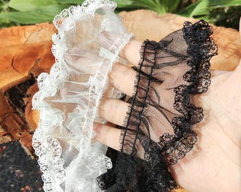Puede incluir: Dos tiras de encaje con volantes, una en blanco y otra en negro. Cada tira presenta una base de malla fina y transparente con un borde decorativo de encaje floral festoneado. La tela es ligera, semitransparente y fruncida para dar volumen.