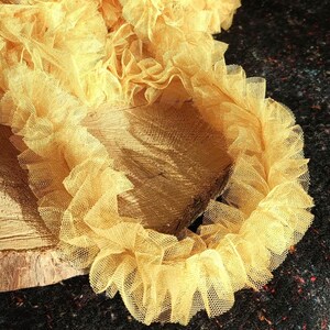Pu&ograve; includere: Un nastro di tulle arricciato color giallo senape con una trama a rete fine. Il tessuto &egrave; arricciato in onde dense e voluminose, creando un aspetto stratificato. La parola Mustard &egrave; stampata in bianco in basso a destra nell'immagine.
