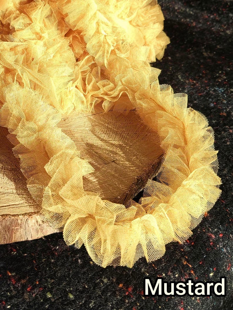 Pu&ograve; includere: Un nastro di tulle arricciato color giallo senape con una trama a rete fine. Il tessuto &egrave; arricciato in onde dense e voluminose, creando un aspetto stratificato. La parola Mustard &egrave; stampata in bianco in basso a destra nell'immagine.