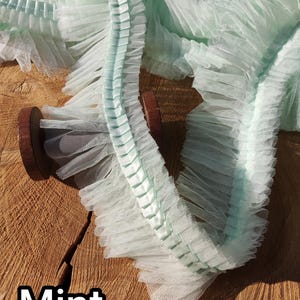 May include: Mint green ruffled tulle trim on a wooden spool. The trim has a delicate, airy texture with a pleated edge. The word "Mint" is displayed in a white, bold font at the bottom of the image.
