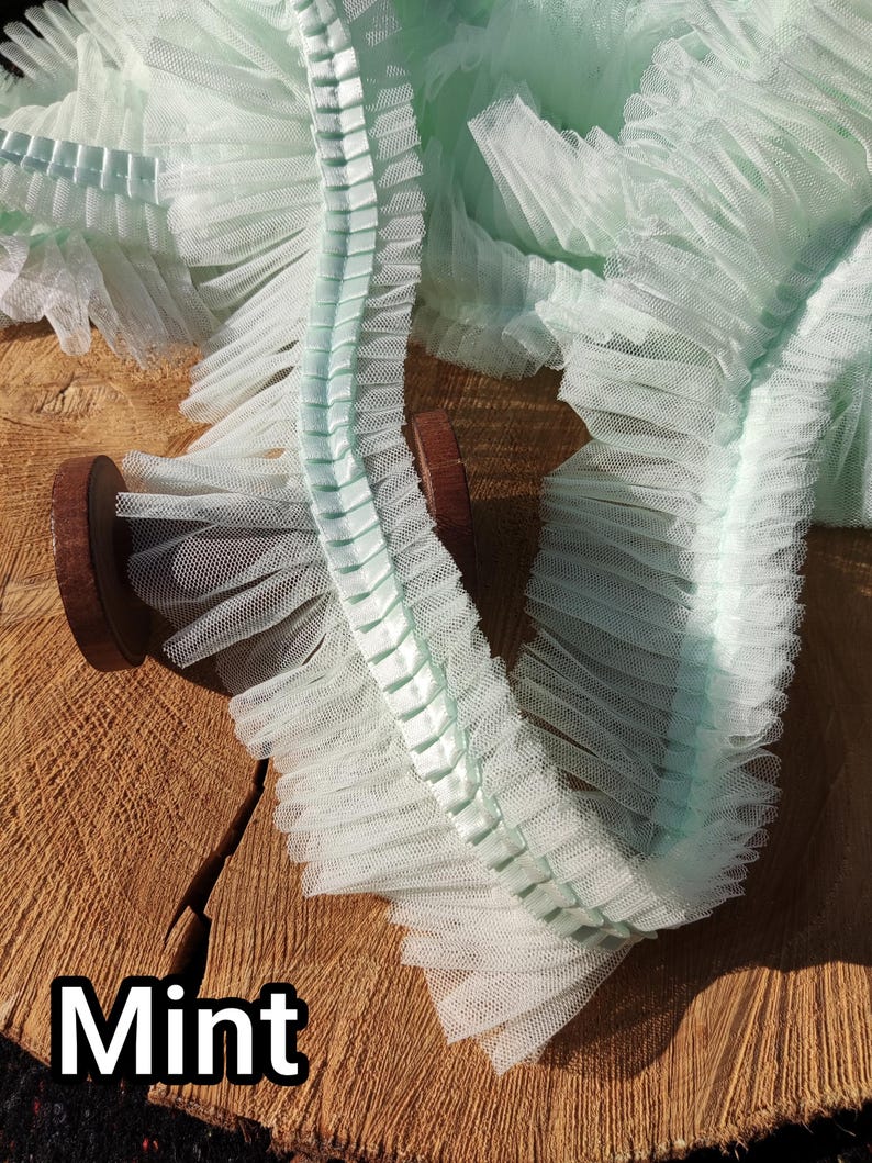 May include: Mint green ruffled tulle trim on a wooden spool. The trim has a delicate, airy texture with a pleated edge. The word "Mint" is displayed in a white, bold font at the bottom of the image.