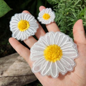Puede incluir: Tres apliques de flores de margarita blancas con centros amarillos. Los apliques son de diferentes tamaños, siendo el más grande el que está en primer plano. Los pétalos están delineados con hilo blanco y los centros son de un amarillo brillante.