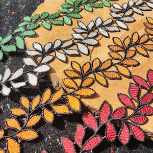May include: Close-up of colourful leaf-shaped embroidered trim. The trim features a black outline with leaves in green, white, orange, and red. The trim is arranged on a wooden surface and a dark, textured fabric.