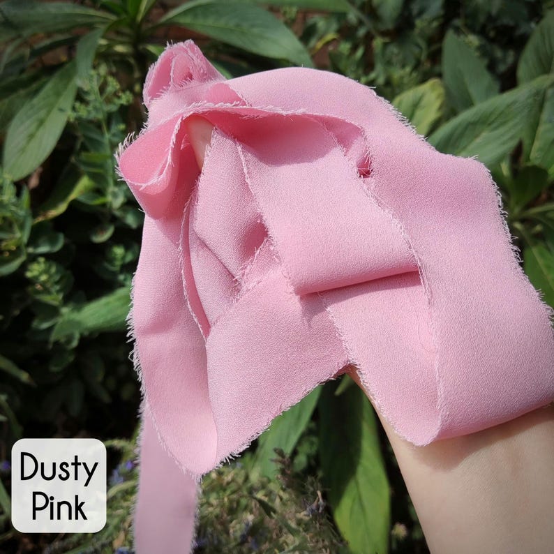 Puede incluir: Una cinta de color rosa polvoriento con bordes deshilachados y sin rematar. La tela tiene una textura suave y mate, drapeada en pliegues sueltos. Una etiqueta cuadrada blanca en la esquina inferior izquierda muestra el texto "Dusty Pink".